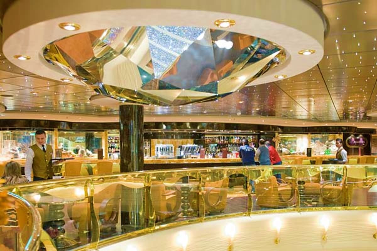 Luxury cruise ship interior atrium with multi-level galleries, gold railings, skylights, and passengers shopping at retail st