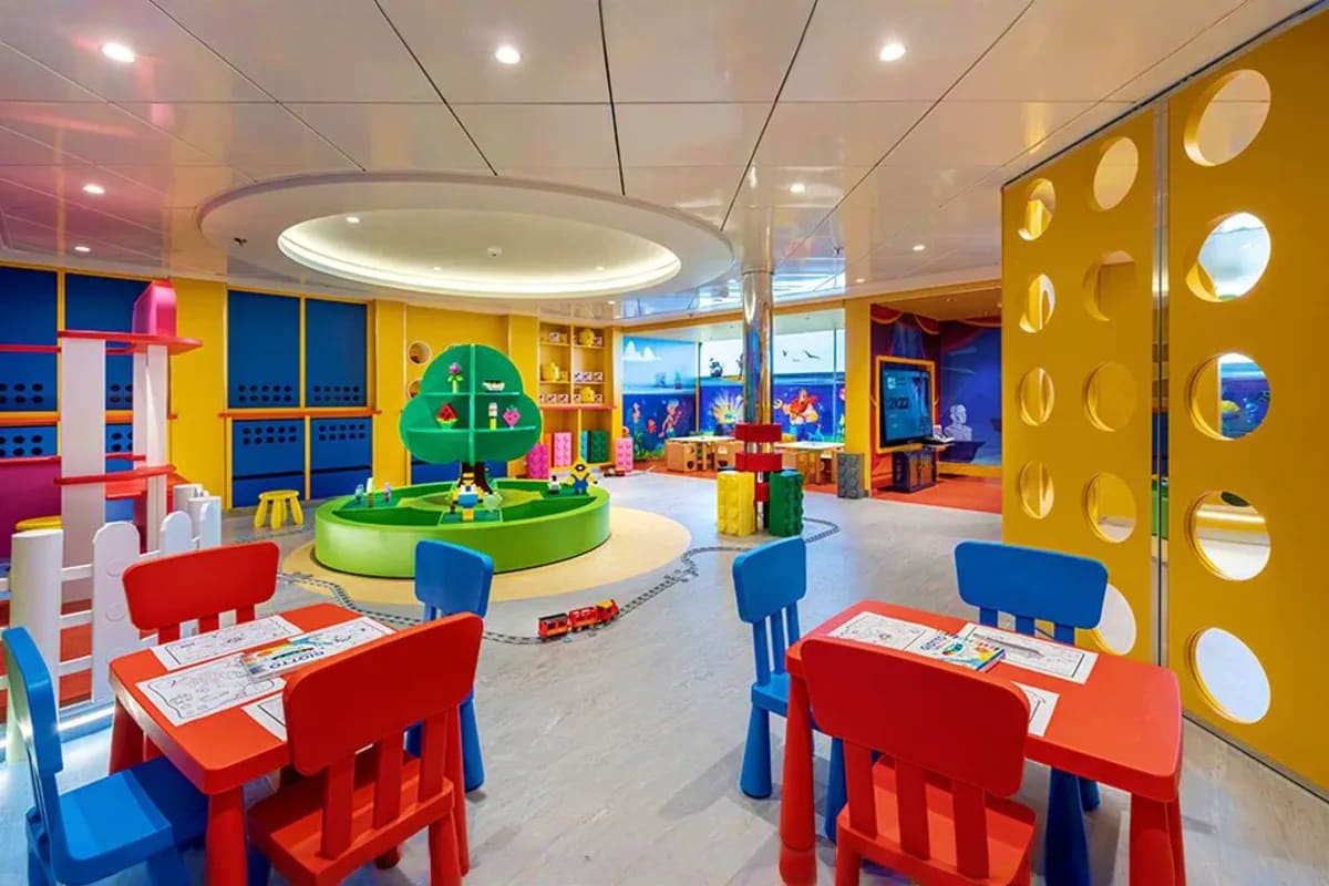 Colorful modern children's play area on cruise ship with activity tables, green play structure, and ocean view windows