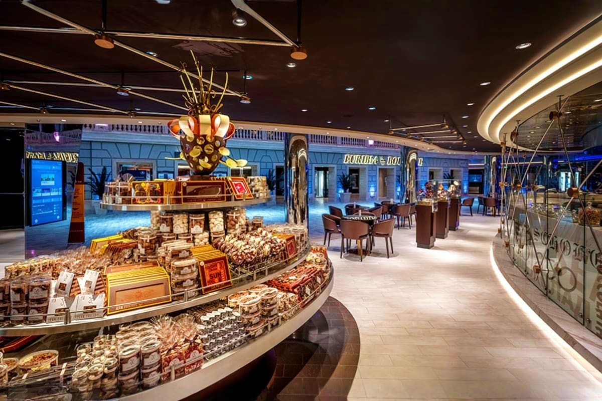 Modern cruise ship specialty food market with curved display counters, packaged goods, and gold crown chandelier centerpiece 