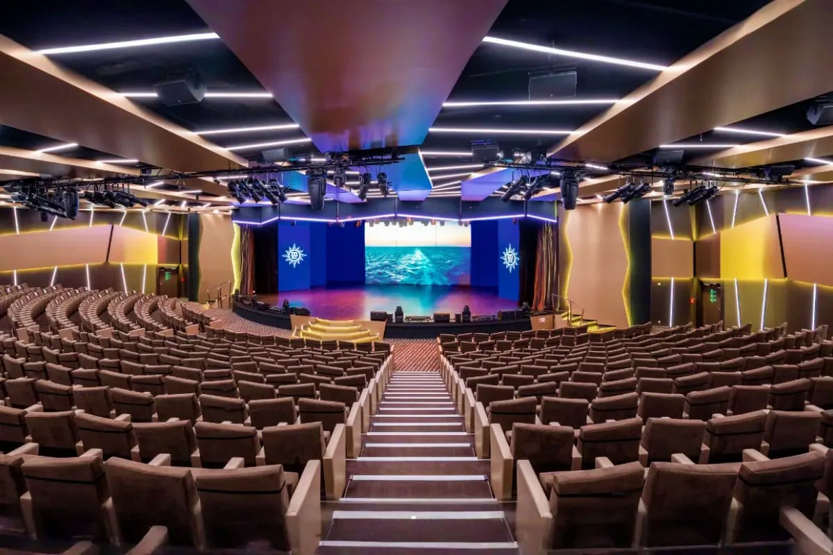 Modern cruise ship theater with rows of beige seats, illuminated stage displaying ocean scenery, and blue nautical decor with