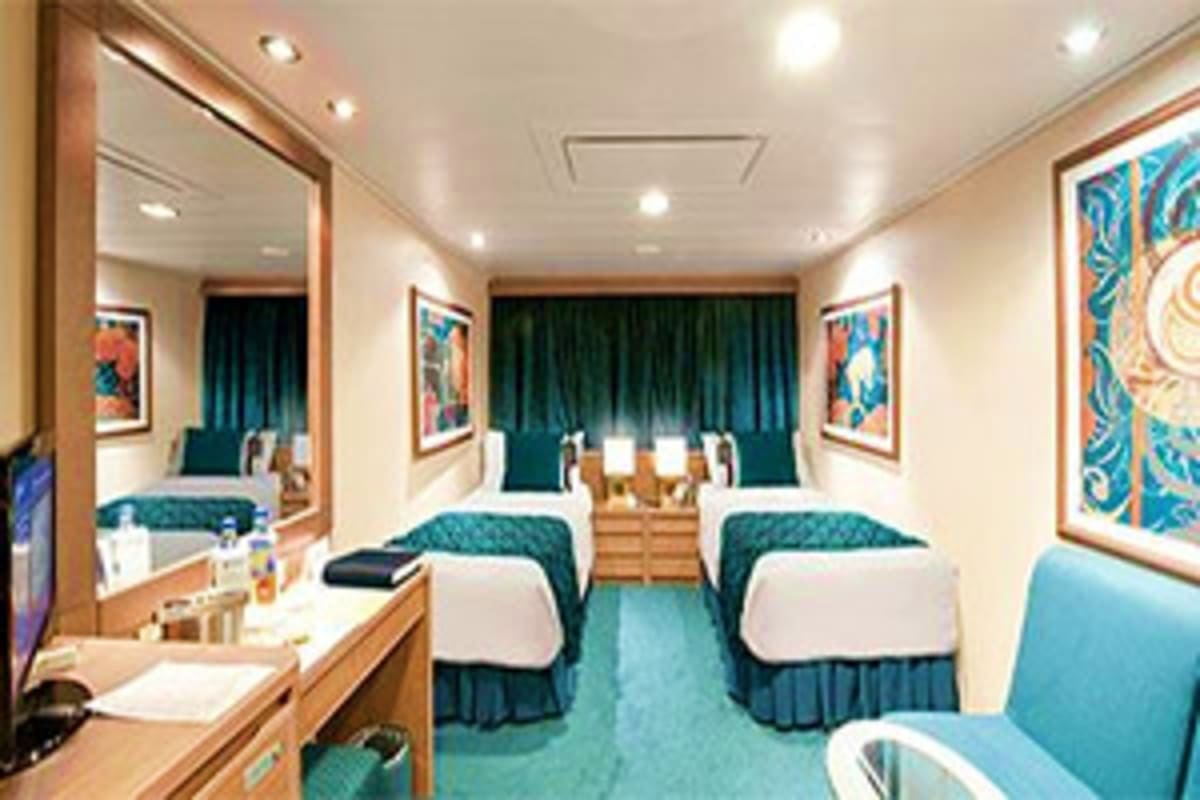 Spacious cruise ship cabin with twin beds, teal accents, wooden furnishings, mirror, and colorful artwork on walls