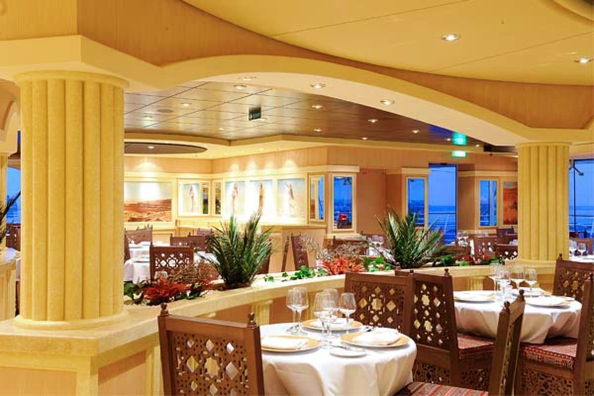 Elegant dining room on MSC Magnifica cruise ship with round tables, white linens, decorative wooden chairs, floral centerpiec