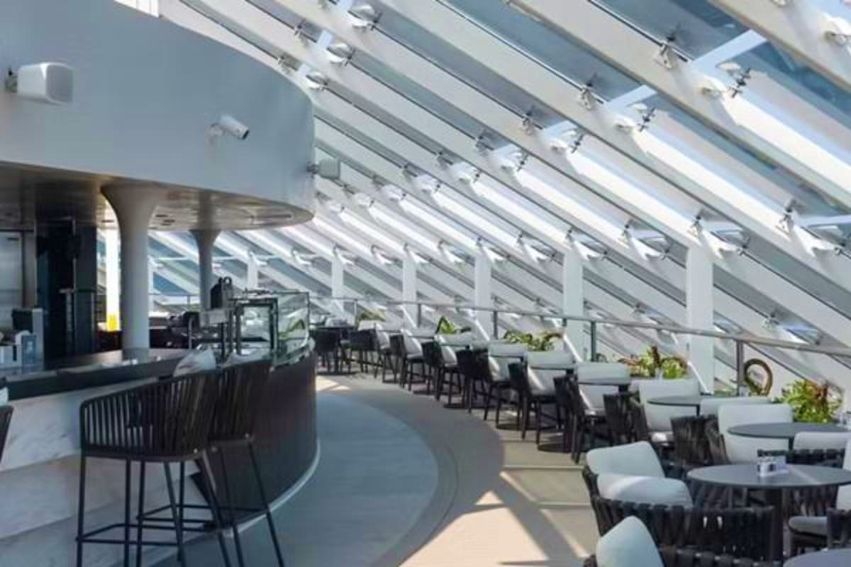 Modern cruise ship indoor dining venue with floor-to-ceiling windows, bar seating, and contemporary furniture overlooking the