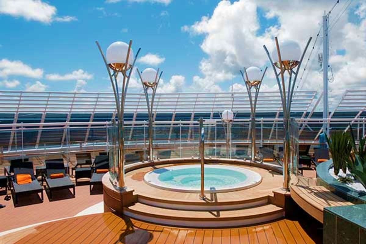 Cruise ship deck with hot tub surrounded by lounge chairs and tall heat lamps under blue sky with clouds