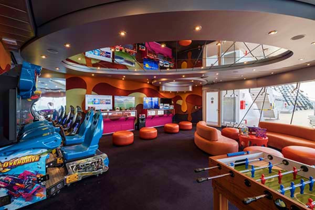 Modern cruise ship entertainment venue with arcade games, orange seating pods, foosball table, and curved ceiling with skylig