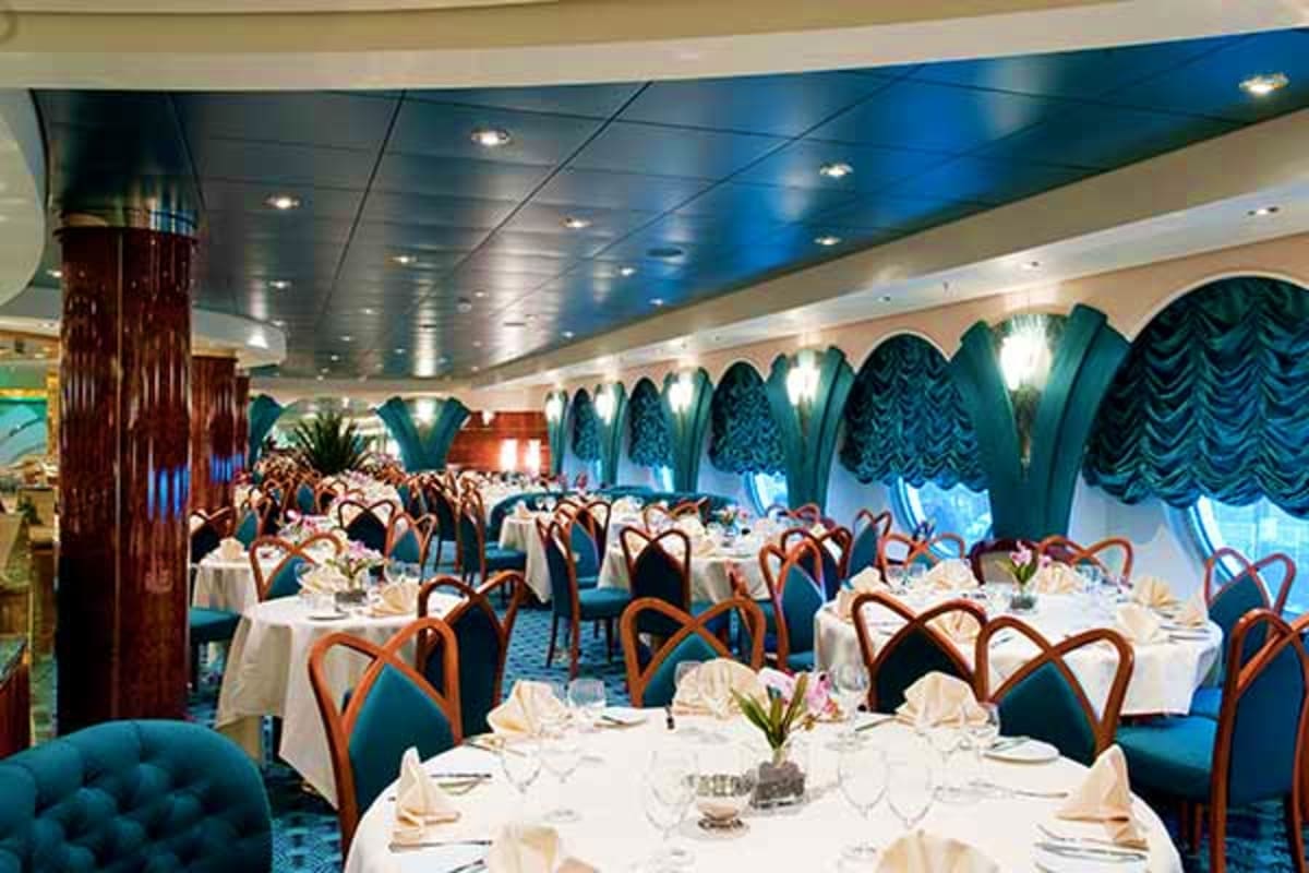 Elegant cruise ship dining room with blue-draped windows, white tablecloths, wooden chairs, and formal place settings