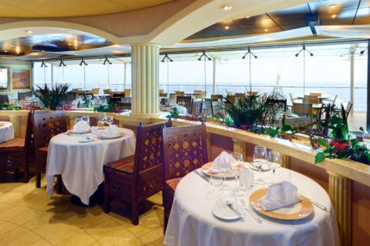 Elegant cruise ship dining room with round tables, white tablecloths, and panoramic ocean views through floor-to-ceiling wind