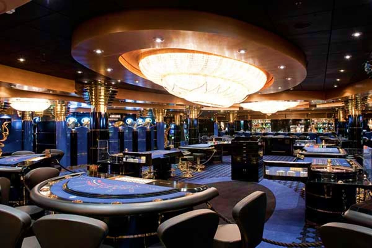 Luxury casino interior on cruise ship with blue carpet, gaming tables, gold accents, and modern lighting fixtures
