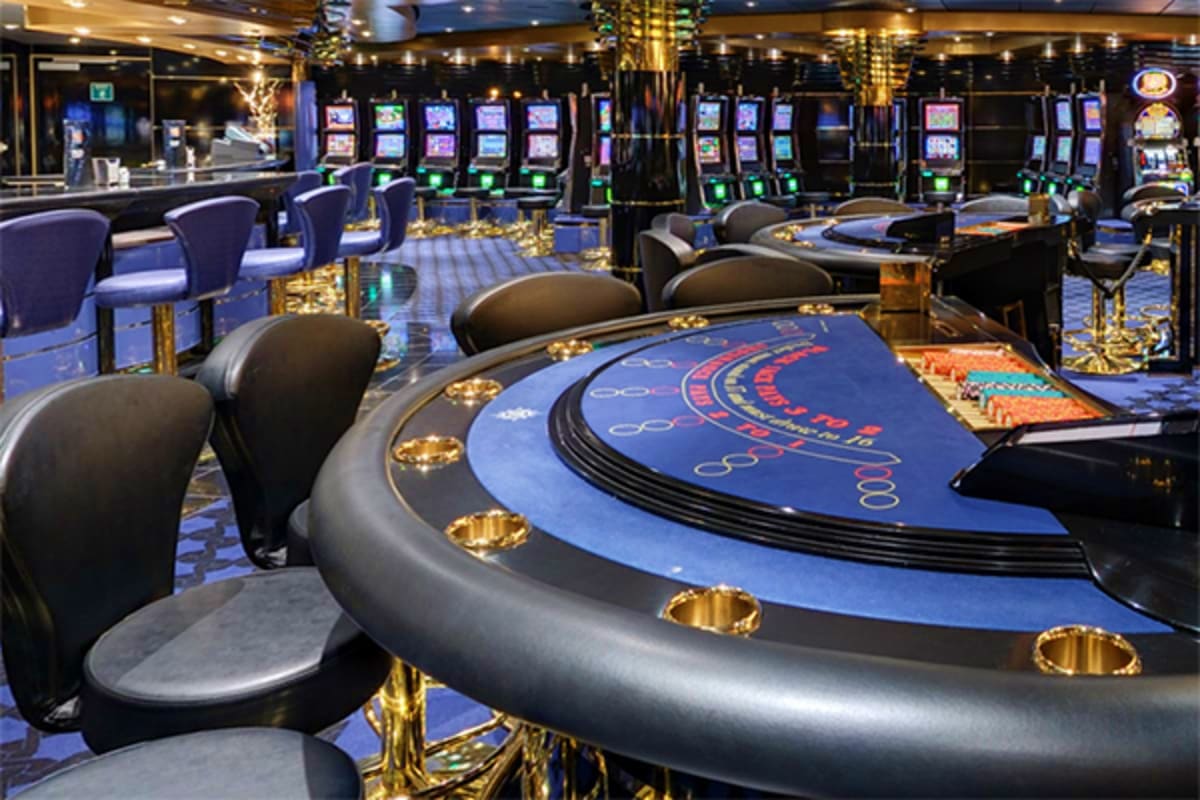 Luxurious casino floor with blue gaming tables, slot machines, and gold accents on a cruise ship