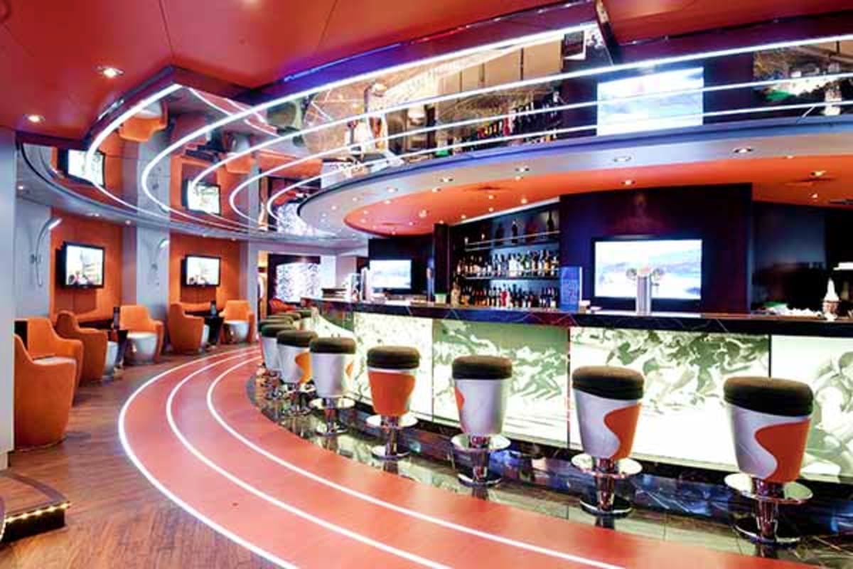 Modern cruise ship interior lounge with orange and blue décor, curved seating areas, bar counter, and multiple screens displa