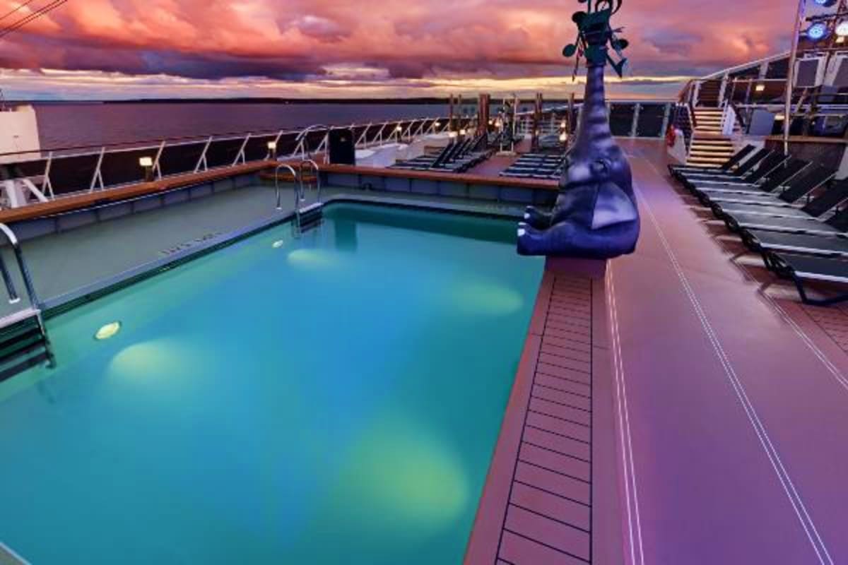 Cruise ship pool deck at sunset with turquoise swimming pool, lounge chairs, and ocean horizon under dramatic pink and purple