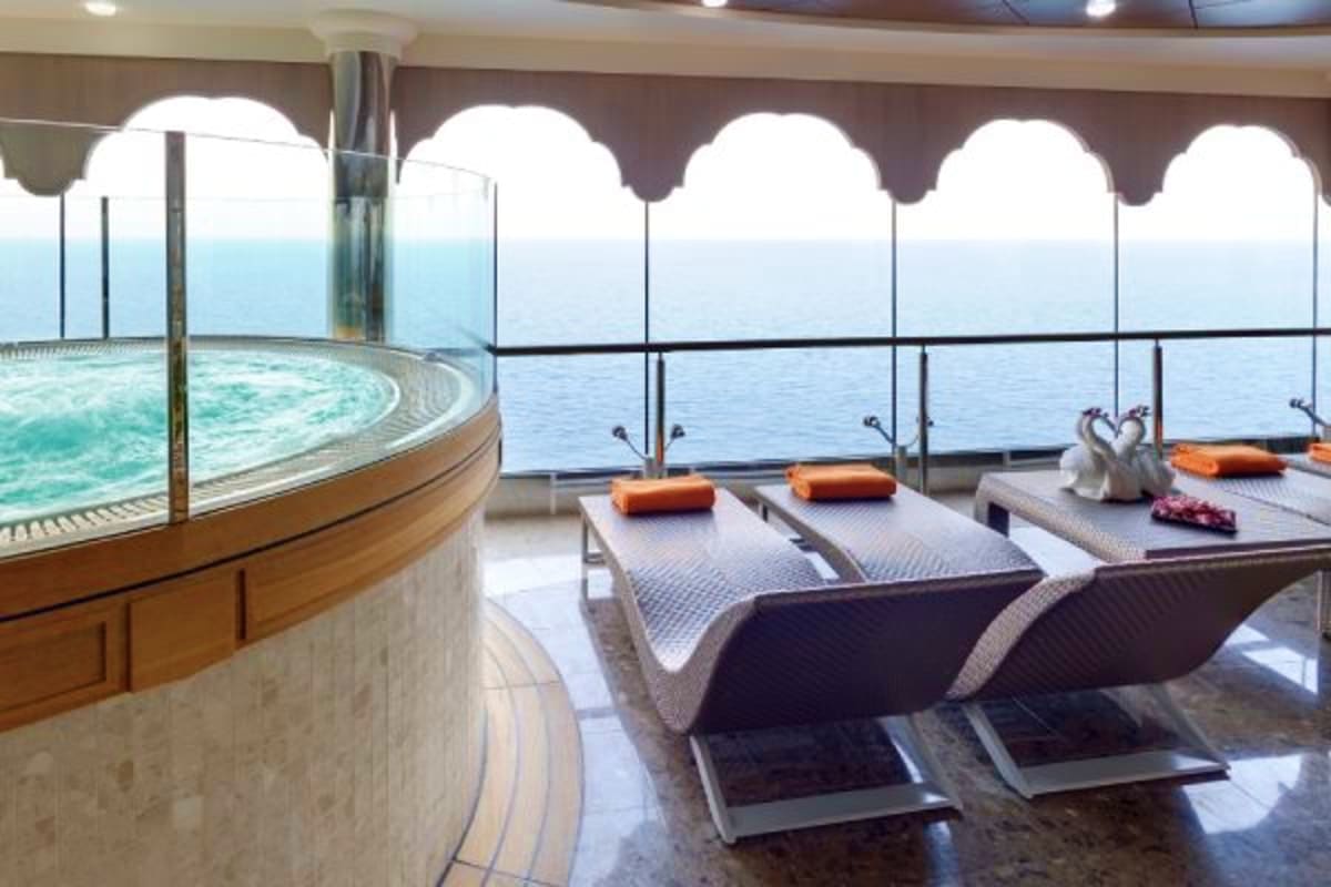 Luxury cruise ship spa deck with hot tub, lounge chairs, and ocean views through floor-to-ceiling windows