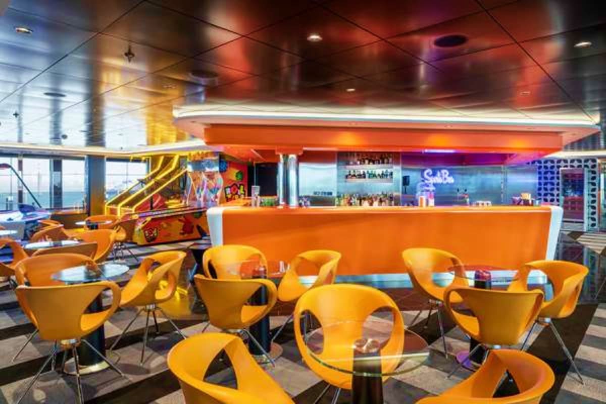 Modern cruise ship lounge with bright yellow chairs, orange bar counter, and colorful neon lighting overlooking the sea