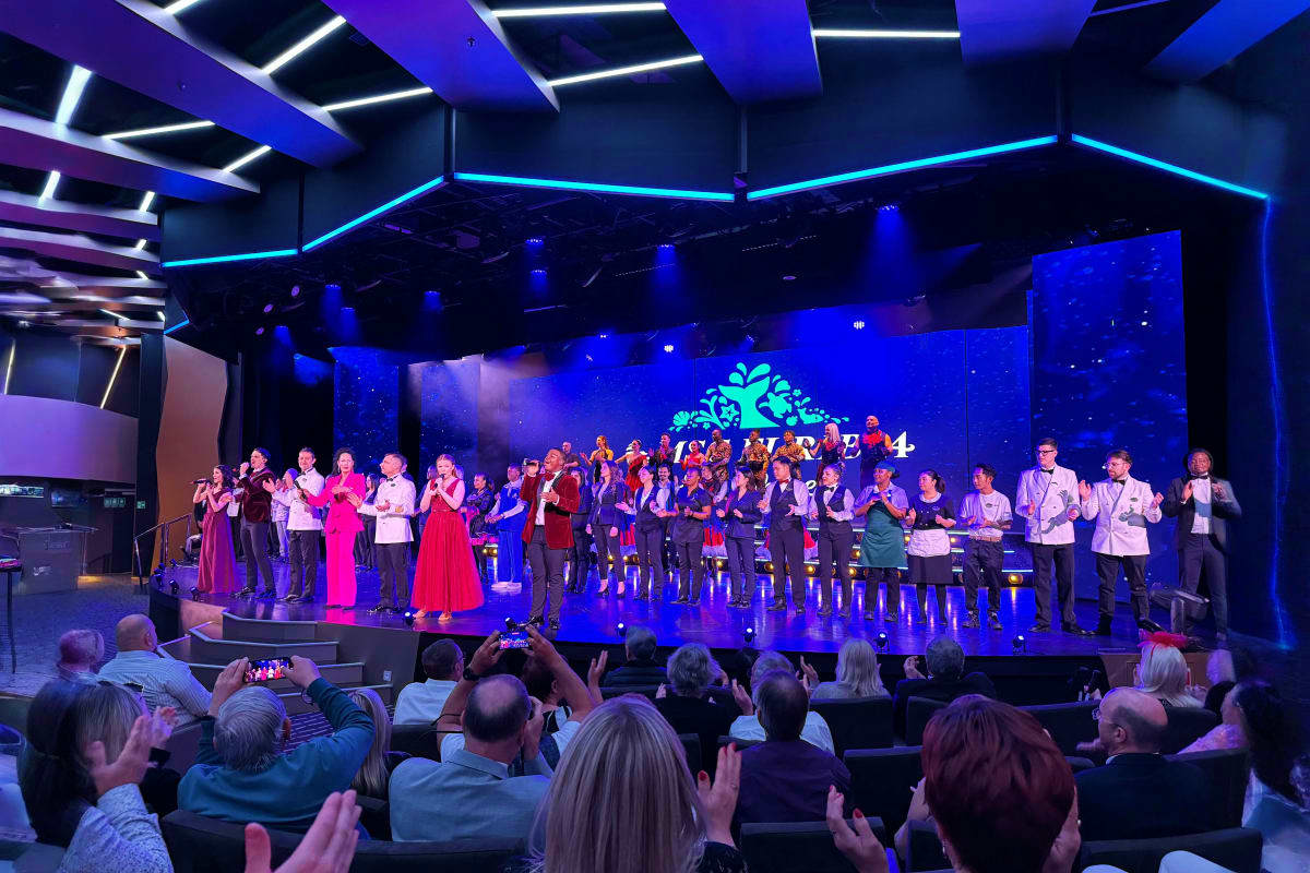 Large choir group performing on illuminated stage with blue and purple lighting while audience members watch and record with 