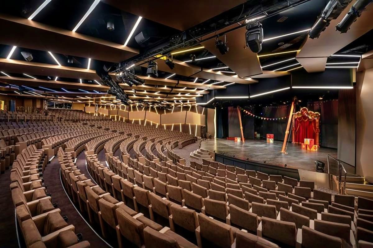 Modern theater auditorium with curved seating rows, stage with red curtains, and contemporary lighting fixtures overhead on M