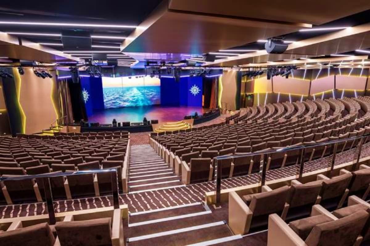 Modern cruise ship theater with rows of beige seats, illuminated stage with ocean video screen, and blue maritime-themed deco