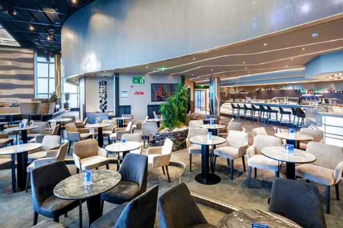 Modern cruise ship lounge with contemporary furniture, bar area, and decorative greenery under ambient lighting