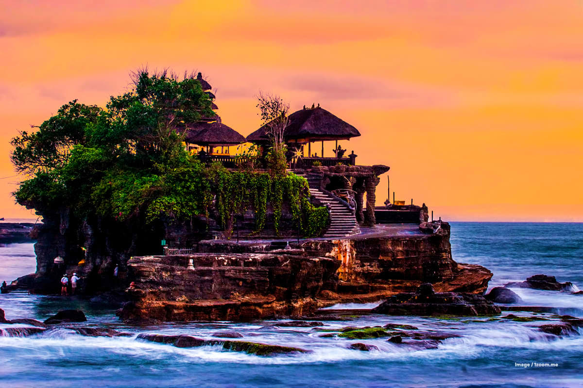 Tanah Lot temple on rocky island at sunset in Bali with traditional thatched-roof buildings and ocean waves in foreground