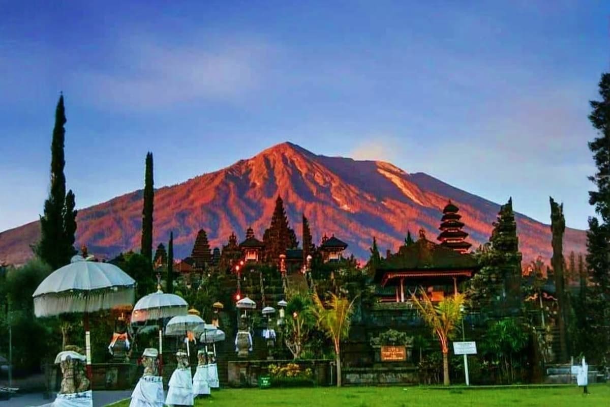 Mount Agung volcano at sunset behind a Balinese temple complex with traditional architecture, cypress trees, and white umbrel