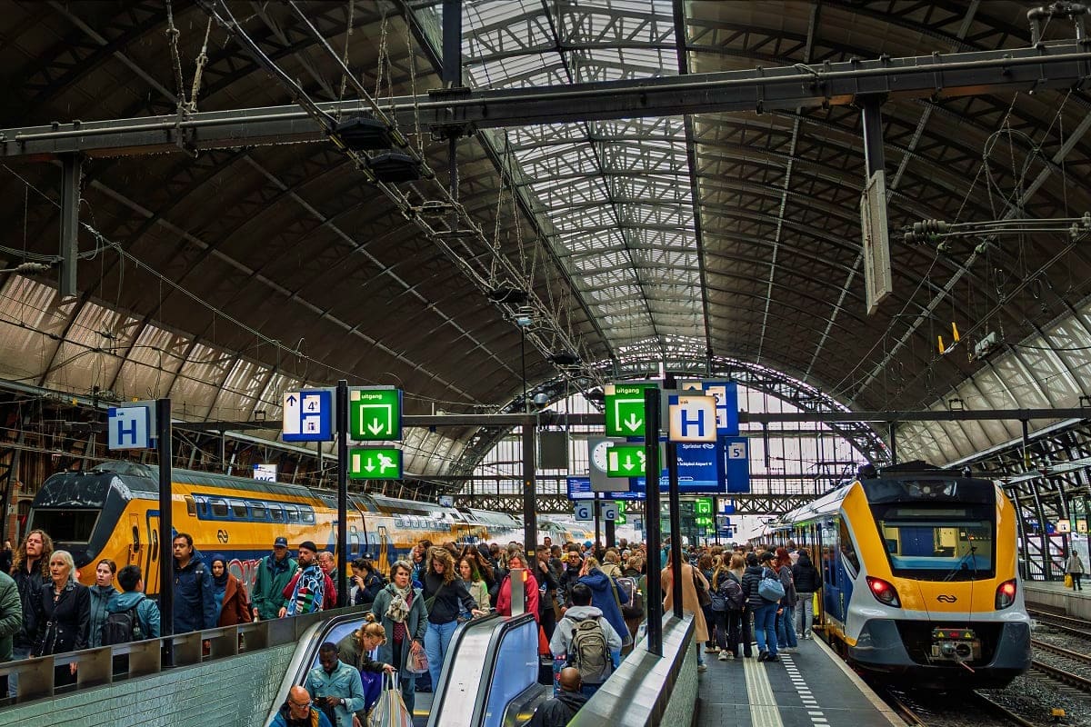 Crowded railway station platform with yellow and blue trains, passengers boarding, metal infrastructure and directional signs