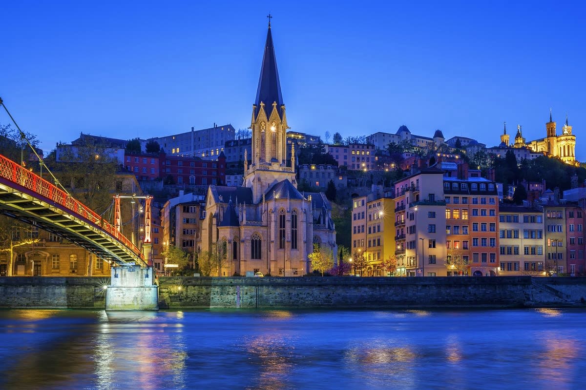 Illuminated Gothic church spire rising above colorful historic buildings along a blue river at twilight in European city