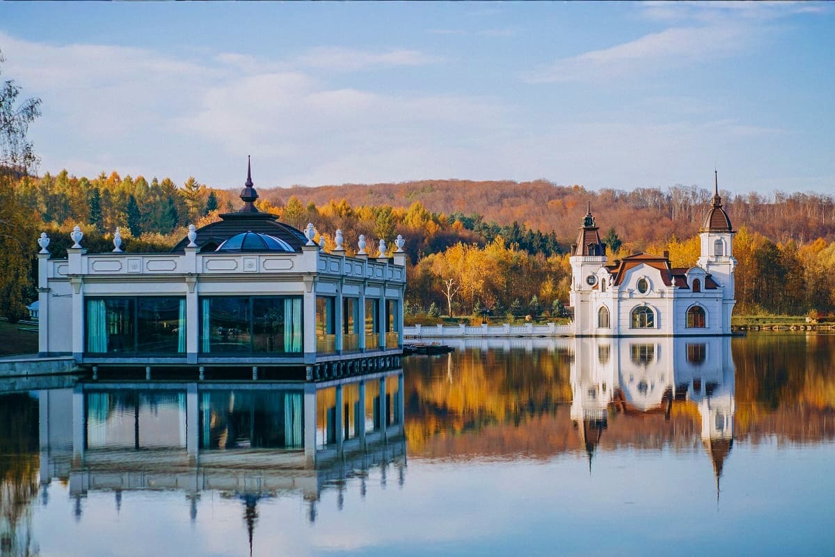 White boathouse and church reflected in calm lake water with autumn forest in background