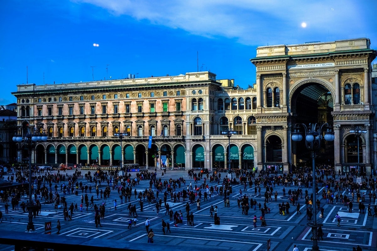 Piazza Duomo in Milan with historic Renaissance buildings, tourists, and ornate architecture under blue sky