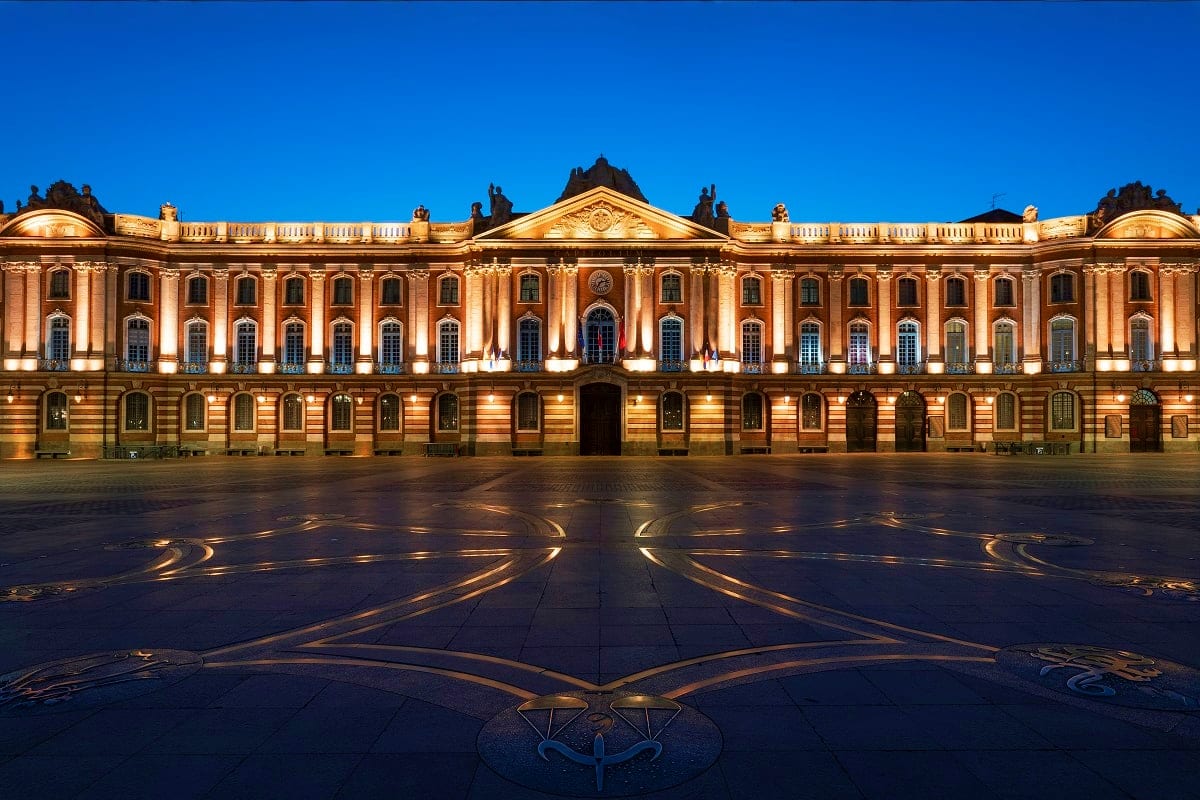 Illuminated neoclassical building at night with golden facade and symmetrical windows overlooking a decorated plaza in Toulou