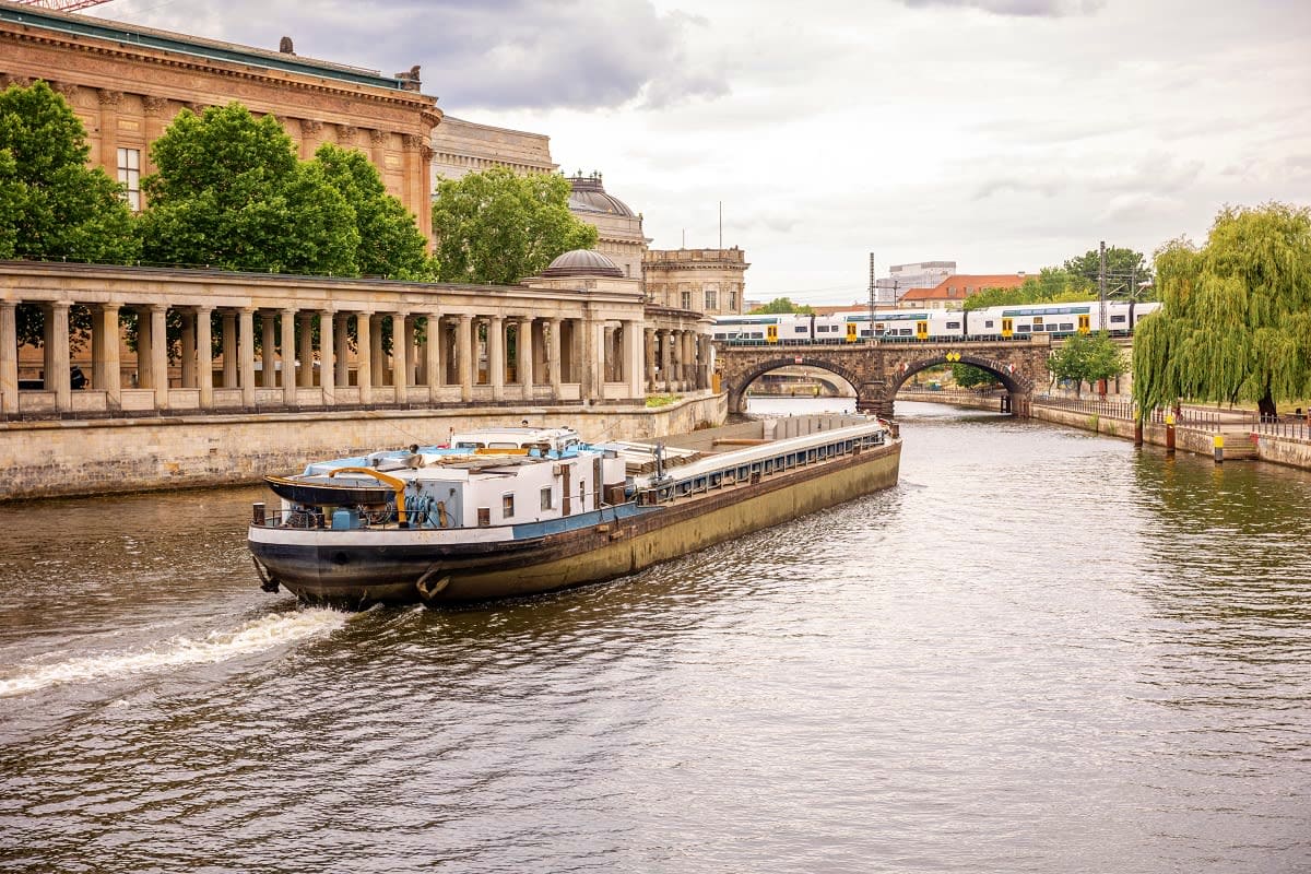 Barge boat on European canal with neoclassical museum building, stone bridge, and passenger train crossing in background