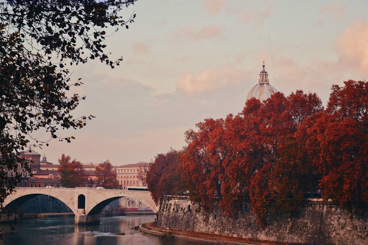 Historic stone bridge over river with autumn foliage and dome-topped basilica in background, Rome Italy cityscape