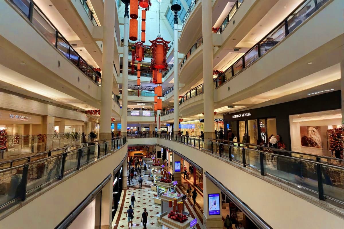 Multi-level shopping mall interior with red lantern decorations and escalators, shoppers browsing retail stores including Bot