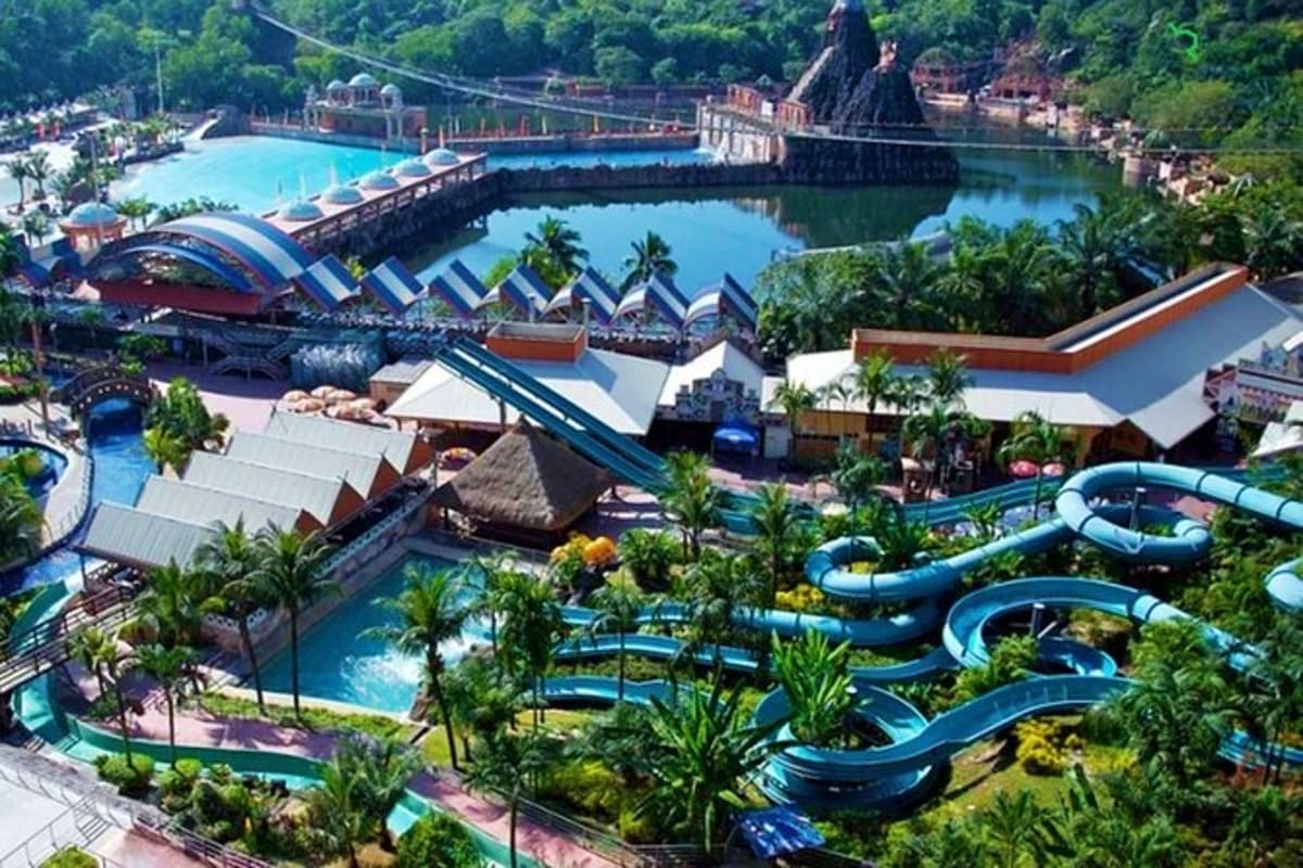 Aerial view of theme park with blue water slides, pools, tropical vegetation, and rock formations surrounded by lush green fo