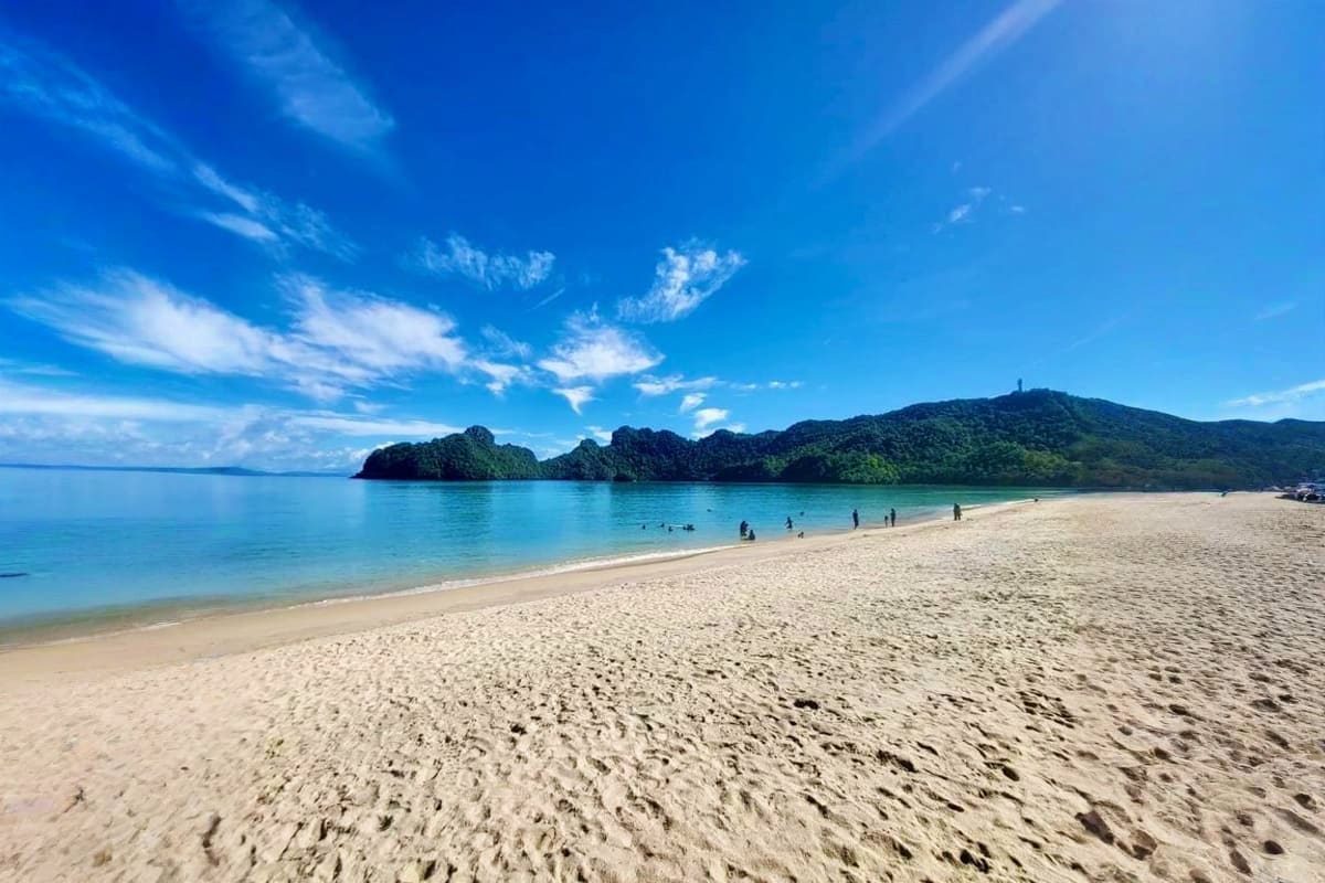 Tropical beach with white sand, turquoise water, and forested islands under blue sky with swimmers in the water