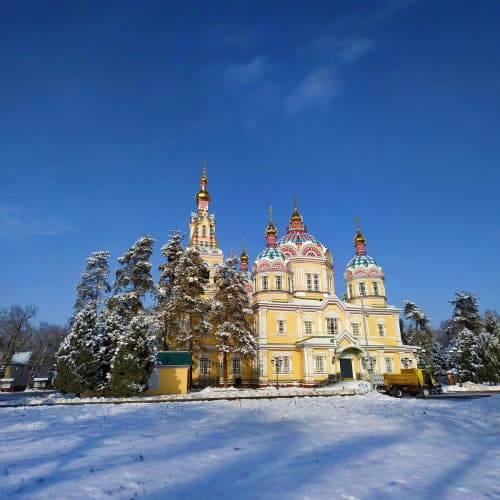 Yellow Orthodox cathedral with golden domes surrounded by snow-covered evergreen trees under clear blue sky in winter