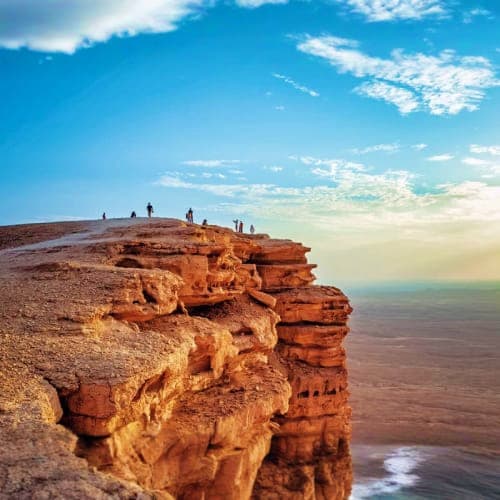 Visitors standing on rocky cliff edge overlooking vast desert landscape under blue sky with white clouds