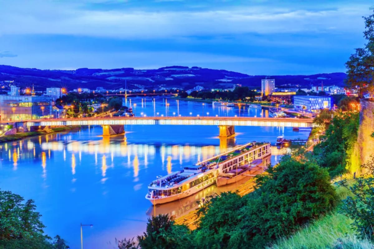 River cruise ship on the Danube River at dusk with illuminated bridge and hillside town in Austria