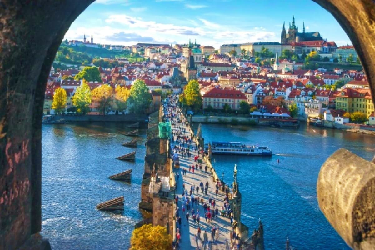 View through stone arch of Charles Bridge spanning Vltava River with Prague cityscape, red-roofed buildings, and castle in ba