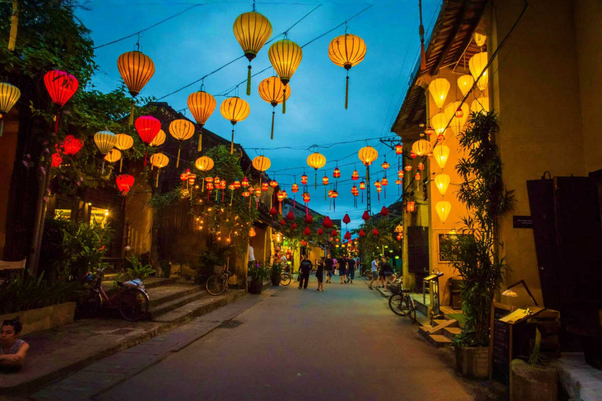 Hoi An ancient town street at dusk with illuminated red and golden lanterns hanging overhead, yellow buildings, and pedestria