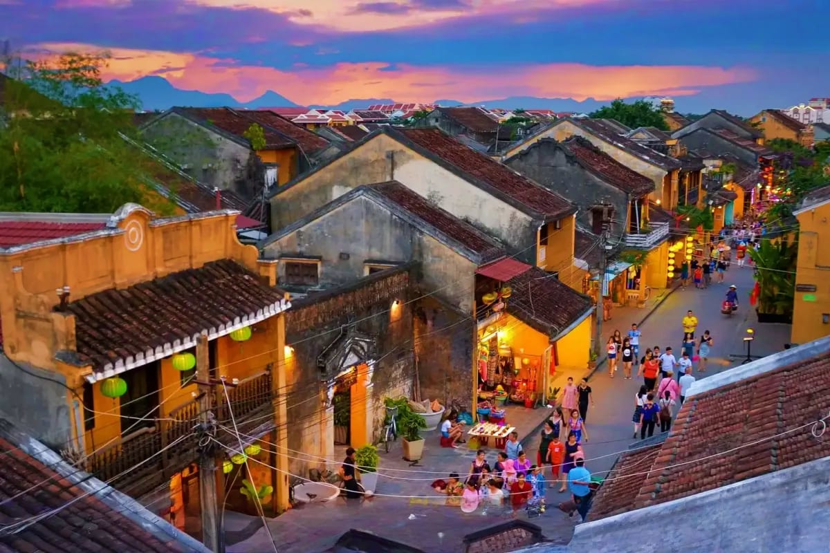 Ancient lantern-lit streets of Hoi An's Old Town at dusk with yellow buildings, tourists, and mountain backdrop at sunset