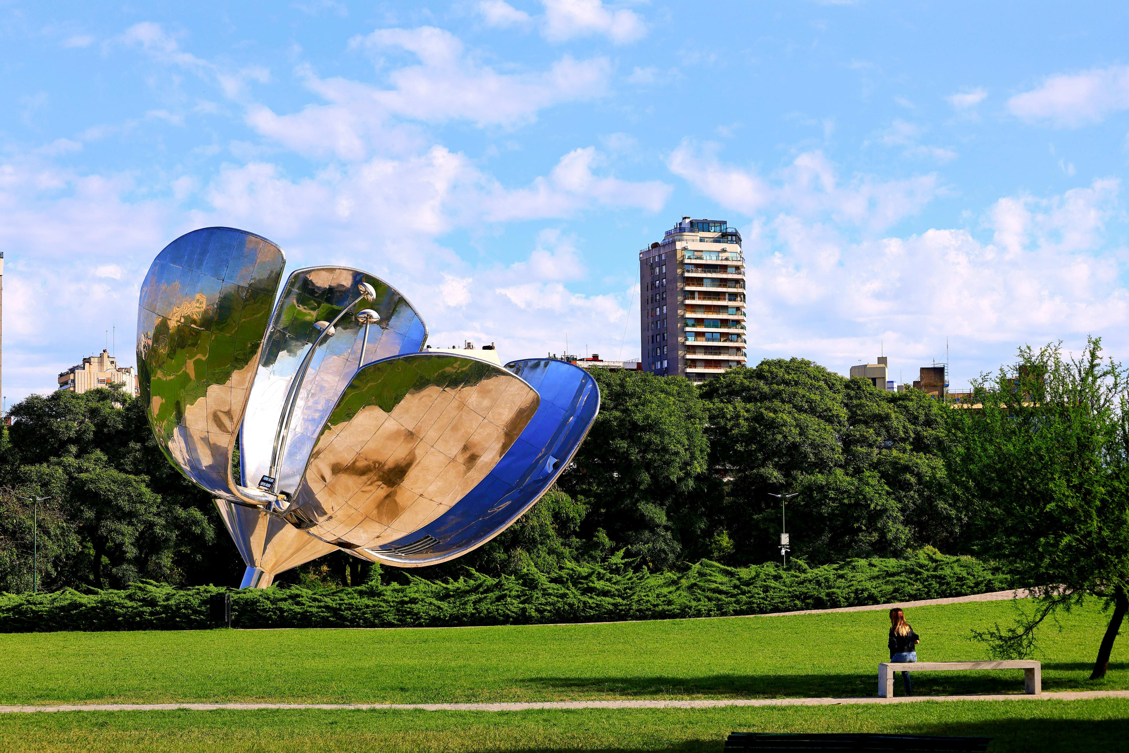 Large reflective metal flower sculpture in urban park with apartment buildings and green trees under blue sky