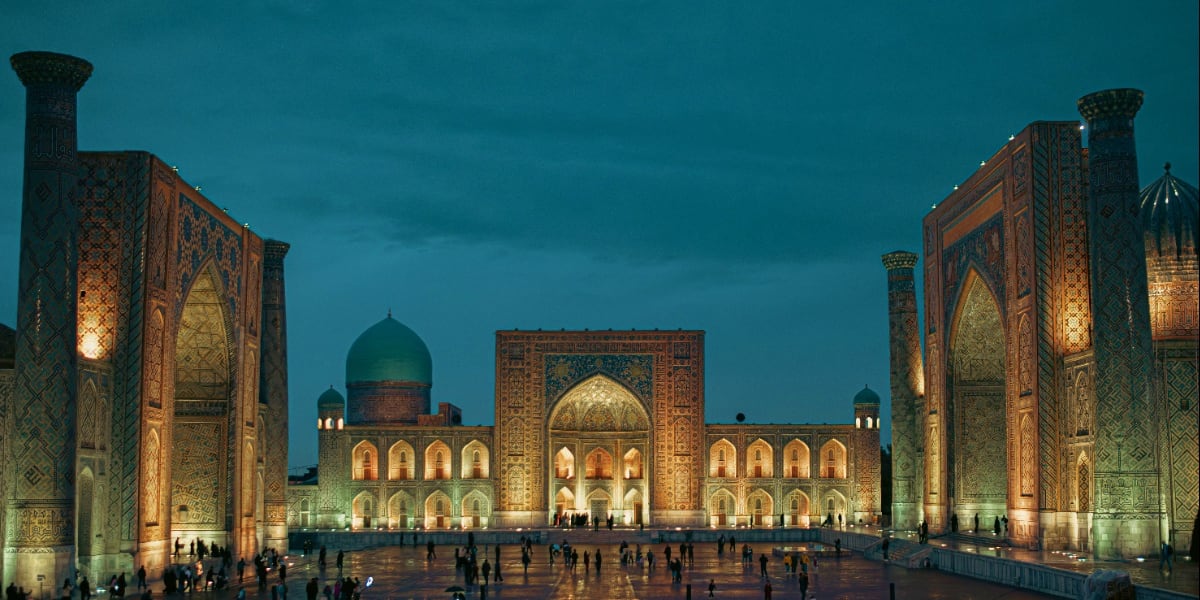 Registan square in Samarkand illuminated at dusk with ornate turquoise domes and decorated facades, visitors gathering in cou