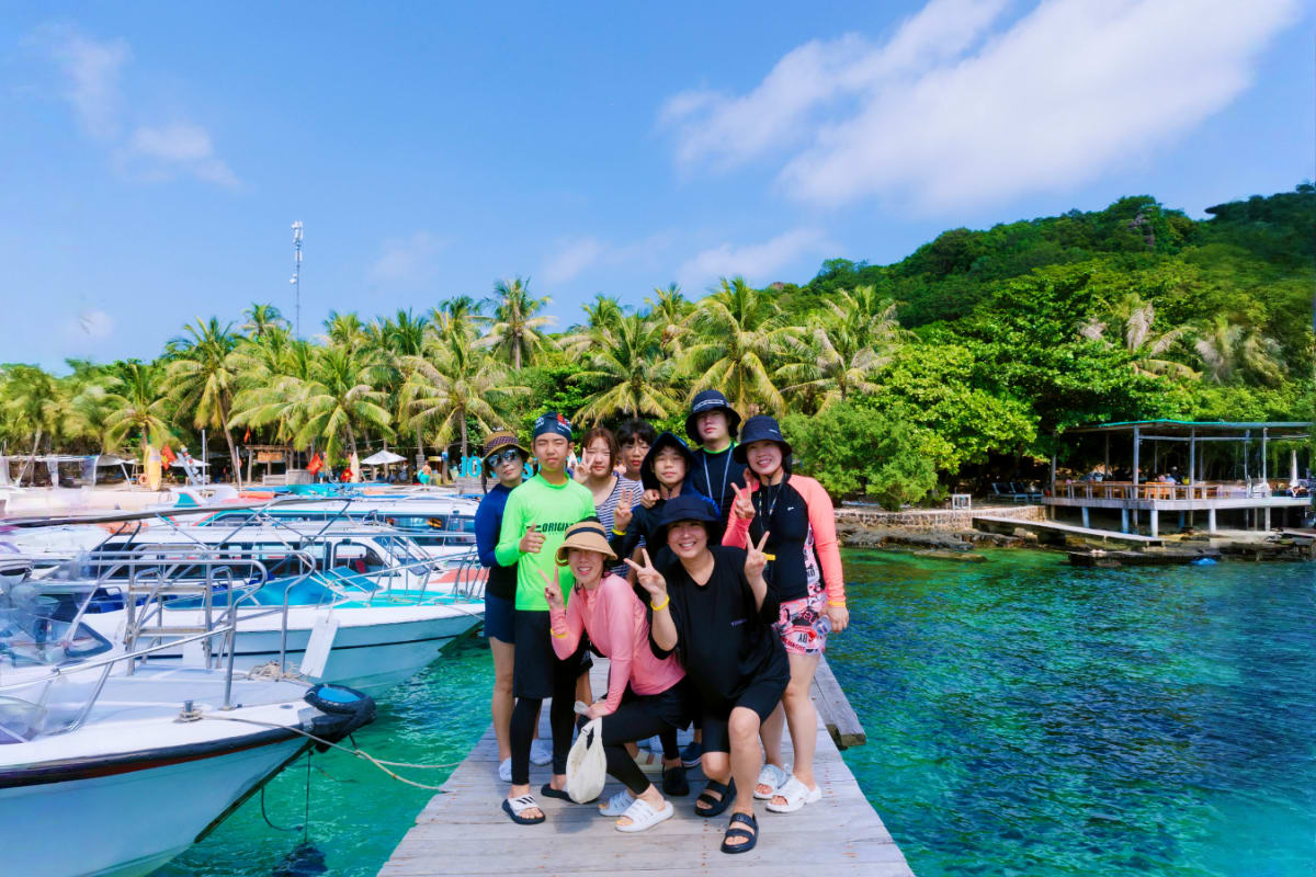 Discovery 3 Island Tour Group of tourists posing on wooden dock at tropical island harbor with moored boats, palm trees, and green hillside in backgr