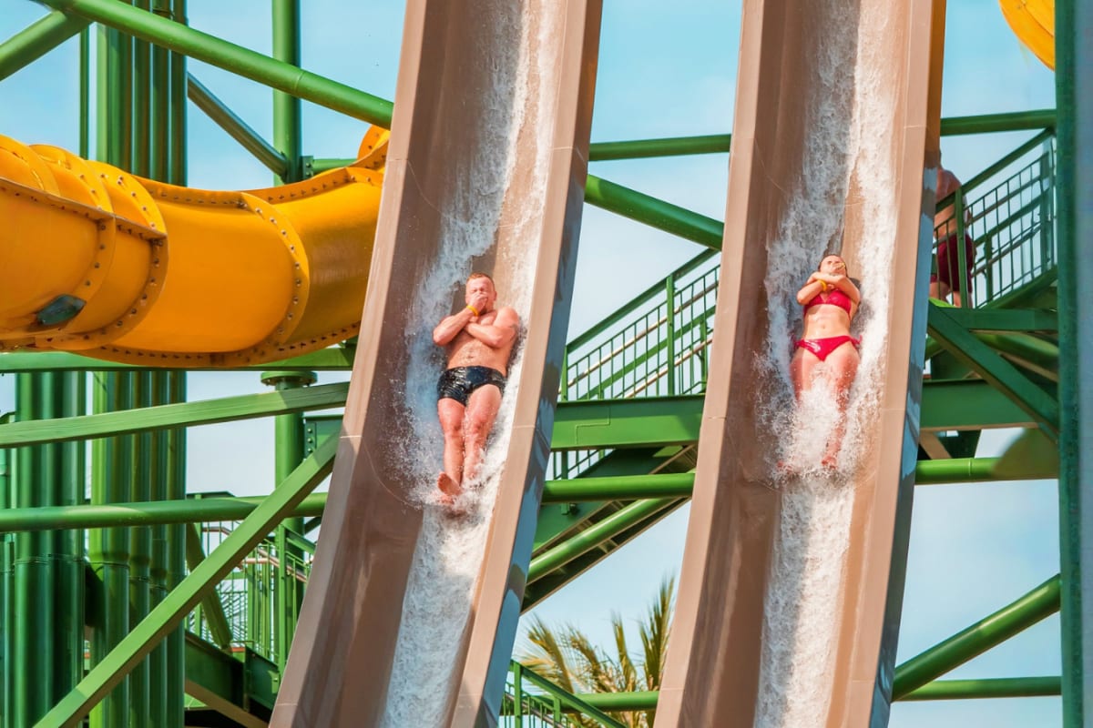 Discovery 3 Island Tour Two people sliding down water slides at a theme park with green metal support structures and yellow pipe visible above