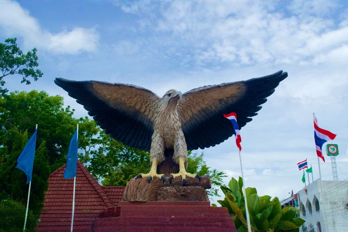 Large eagle monument statue with spread wings in Krabi city square surrounded by Thai flags and green trees under blue sky
