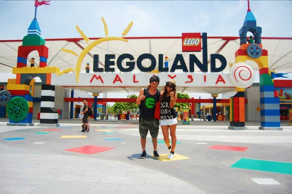 Two visitors posing in front of the colorful LEGOLAND Malaysia entrance sign with LEGO brick sculptures and towers on either 