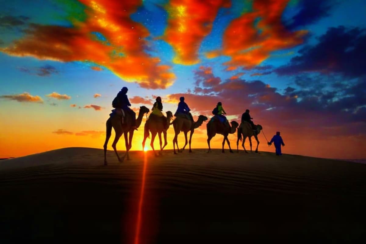 Camel Riding Silhouetted caravan of tourists riding camels across sand dunes at sunset with vibrant orange and blue sky