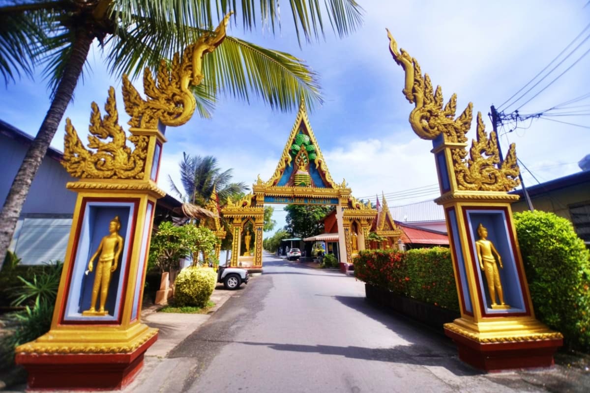 Ornate golden Thai temple gates with Buddha statues flanking entrance road leading to central temple building in Koh Samui