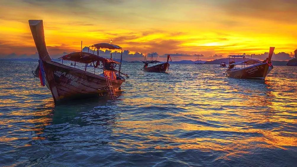 7 Island tour by Long Tail Boat Traditional long tail boats anchored in shallow water at golden sunset with mountains in background