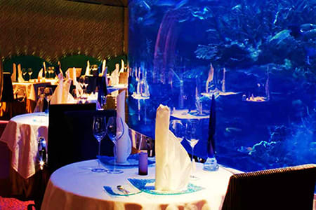 Burj Al Arab Afternoon Tea Elegant dining table set with white linens and place settings overlooking massive blue aquarium with swimming fish at luxury
