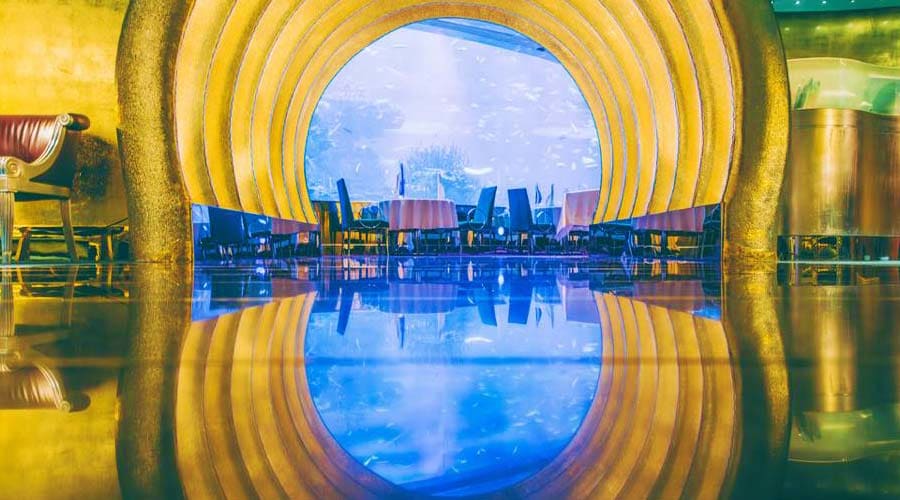 View through yellow industrial pipe tunnel to Dubai waterfront with Burj Al Arab hotel and boats reflected in still blue wate